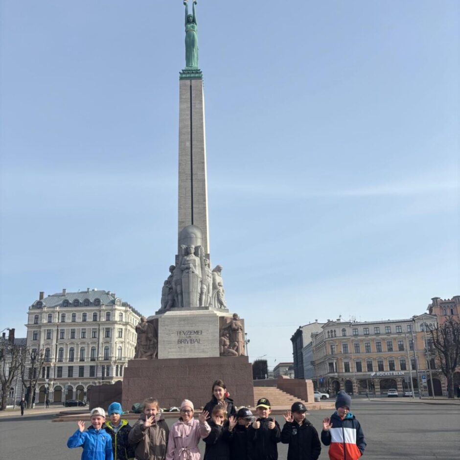 Desmit bērni stāv rindā Rīgas Brīvības pieminekļa priekšā, daži māj ar roku. Aizmugurē redzams piemineklis ar uzrakstu 
