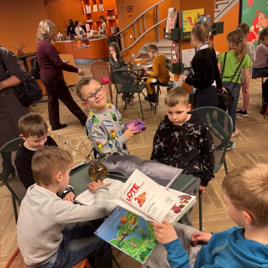 Vairāki bērni sēž pie galdiem bibliotēkā vai grāmatnīcas bērnu stūrī. Priekšplānā zēns ar brillēm smaida kamerā, turot purpursarkanu kastīti. Viņam blakus un pretī citi zēni lasa grāmatas. Uz vienas grāmatas vāka skaidri redzams teksts 