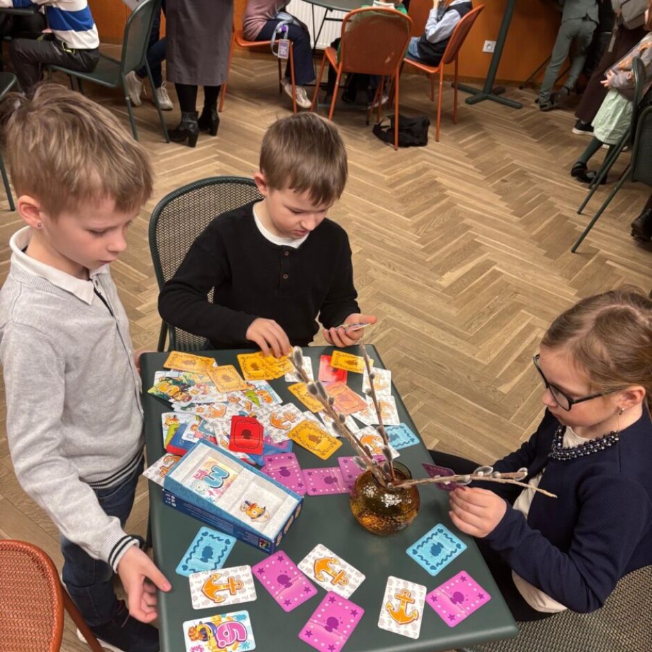 Trīs bērni spēlē galda spēli pie zaļa galda ar kartītēm. Divi zēni skatās uz spēļu kārtīm, bet meitene ar brillēm tur vāzē ieliktus pūpolzarus. Uz spēles kastes ir uzraksts 