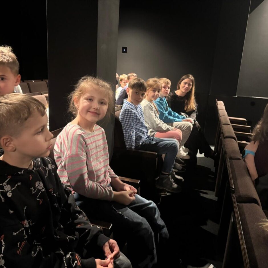 Bērnu un pieaugušo grupa sēž tumšā auditorijā ar sēdvietu rindām, skatoties uz priekšu. Priekšplānā meitene ar svītrainu džemperi smaida kamerā.