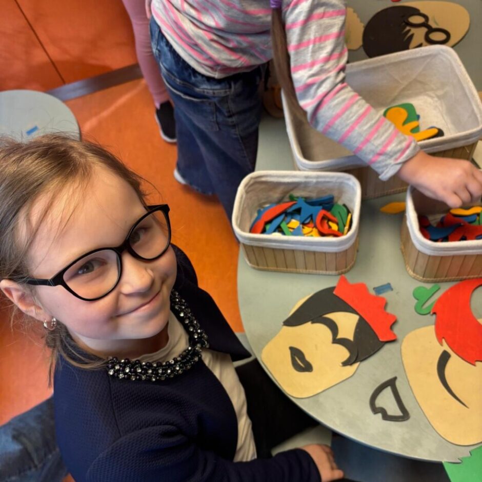 Jauna meitene brillēs, tumši zilā žaketē ar melnu kaklarotu, smaida un skatās uz kameru, sēžot pie galda. Uz galda ir konteineri ar krāsainiem filca gabaliņiem un koka figūrām, pie kurām meitenes veido sejas. Fona redzama cita bērna roka, kas darbojas ar materiāliem.