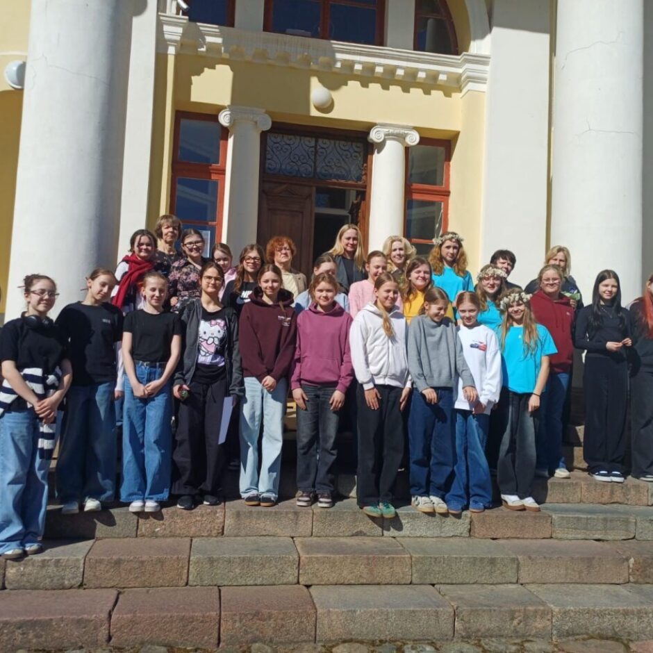 Liela grupa cilvēku, galvenokārt jaunas sievietes un meitenes, pozē fotogrāfijai uz akmens pakāpieniem pie gaiši dzeltenas ēkas ar baltām kolonnām.