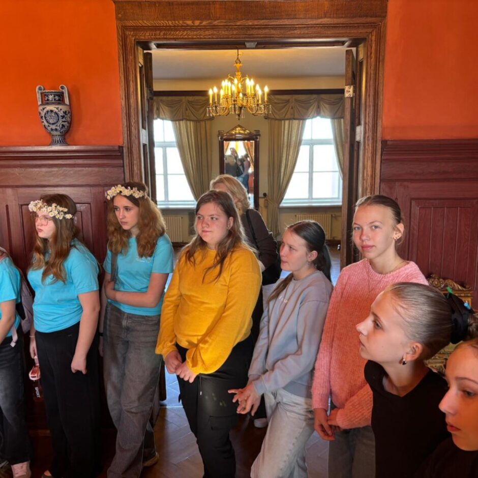 Jaunu meiteņu grupa stāv greznā telpā ar oranžām sienām un koka apdari. Dažām meitenēm galvās ir ziedu vainagi. Fonā caur durvīm redzama lustra un logs.