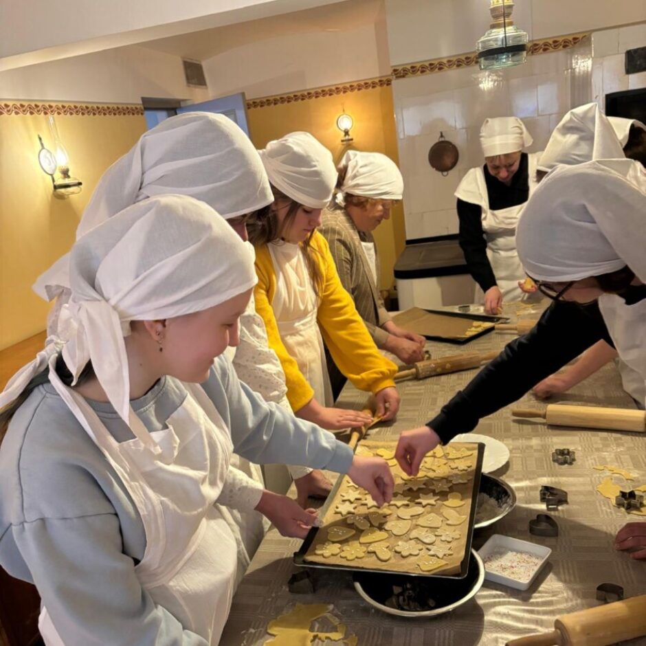 Grupa cilvēku, galvenokārt jaunas sievietes baltos priekšautos un lakatiņos, ap galdu cep sīkdatnes. Viena persona kārto izgrieztās mīklas formas uz cepešpannas, kamēr cita pārkaisa tām virsmu. Uz galda redzami vairāki koka rullīši un cepumu formiņas blakus izrullētai mīklai.