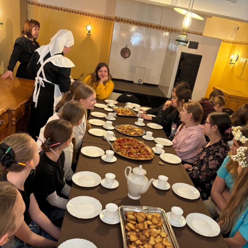 Grupa cilvēku, galvenokārt jaunas meitenes, sēž pie gara galda senu laiku istabā. Galds klāts ar baltiem šķīvjiem, tasītēm, tējkannu un ēdieniem, tostarp pīrāgu un paplāti ar smalkmaizītēm. Pie galda gala stāv divas sievietes, viena no tām tērpusies melnā kalpones uniformā ar baltu priekšautu un galvassegu.