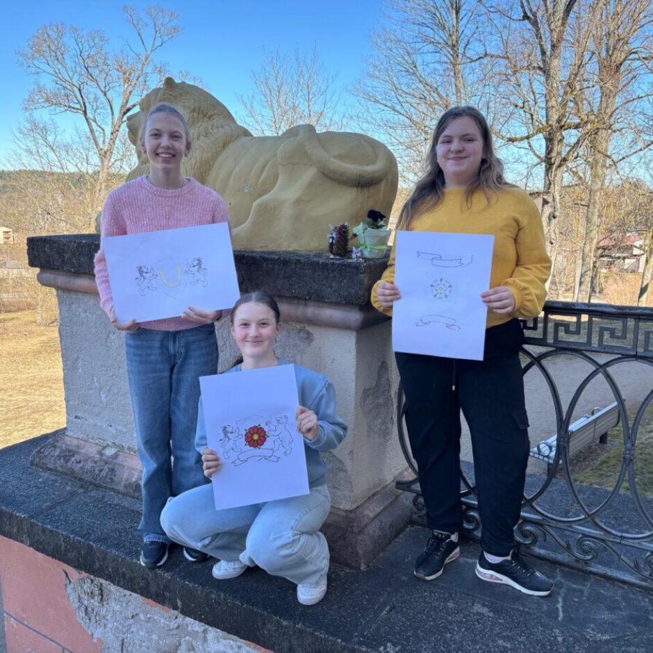 Trīs smaidīgas meitenes ārā tur rokās zīmējumus ar lentu un ziedu motīviem. Aiz viņām redzama zelta lauvas statuja, koki un tāla ainava saulainā dienā.