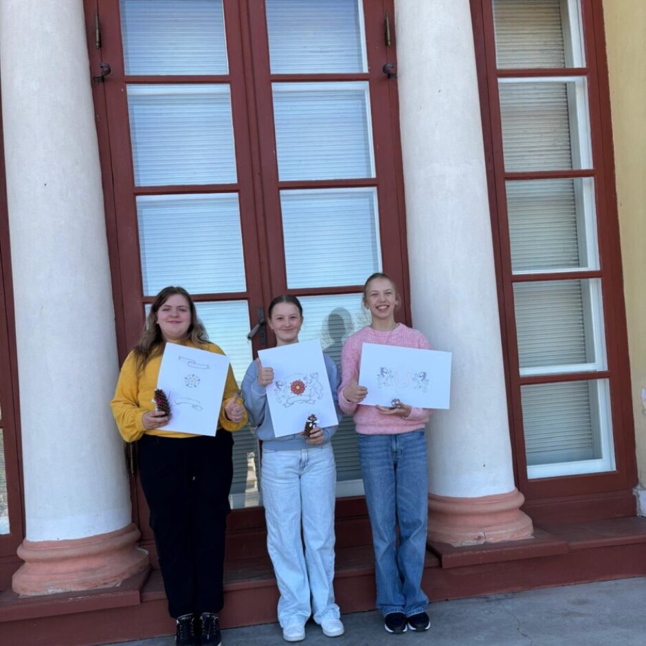 Trīs smaidīgas meitenes stāv pie ēkas ar augstiem logiem un kolonnām, turot rokās krāsainus zīmējumus, un divām no viņām ir arī nelielas balvas.