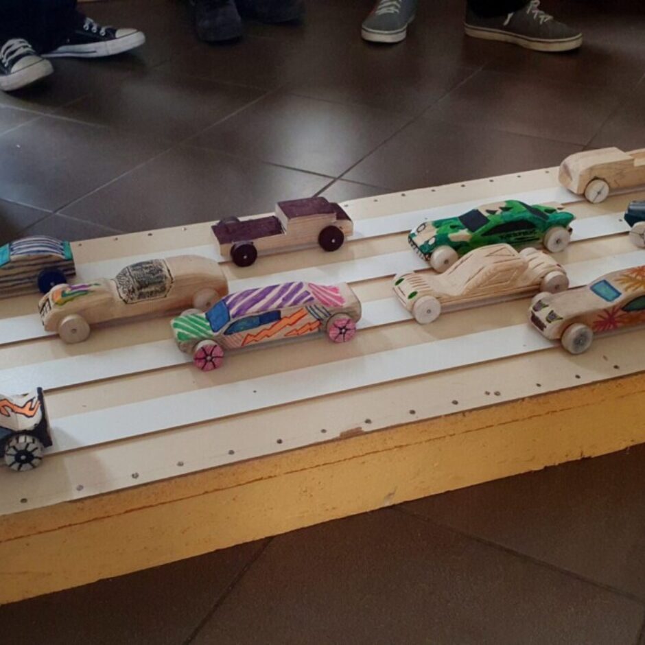 Krāsainas, paštaisītas koka sacīkšu automašīnas dažādos dizainos sarindotas uz daudzjoslu koka trases, gatavas sacīkstēm. Fonā redzamas vairākas cilvēku kājas.