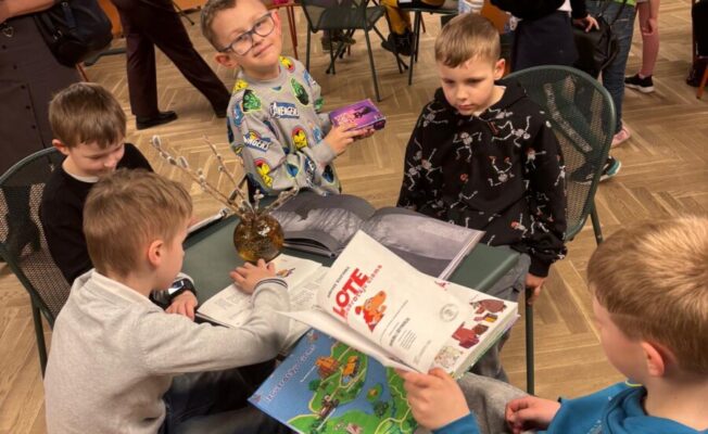 Vairāki bērni sēž pie galdiem bibliotēkā vai grāmatnīcas bērnu stūrī. Priekšplānā zēns ar brillēm smaida kamerā, turot purpursarkanu kastīti. Viņam blakus un pretī citi zēni lasa grāmatas. Uz vienas grāmatas vāka skaidri redzams teksts 