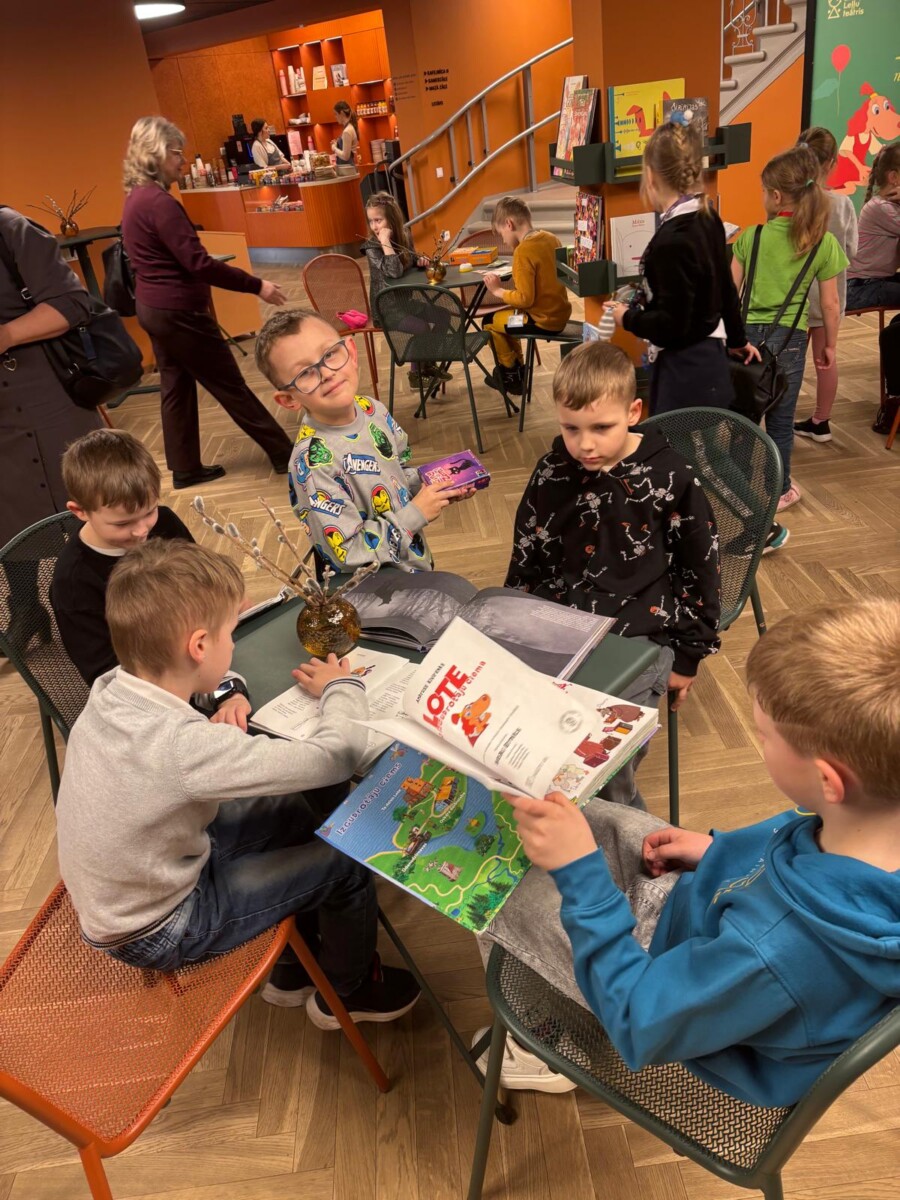 Vairāki bērni sēž pie galdiem bibliotēkā vai grāmatnīcas bērnu stūrī. Priekšplānā zēns ar brillēm smaida kamerā, turot purpursarkanu kastīti. Viņam blakus un pretī citi zēni lasa grāmatas. Uz vienas grāmatas vāka skaidri redzams teksts 