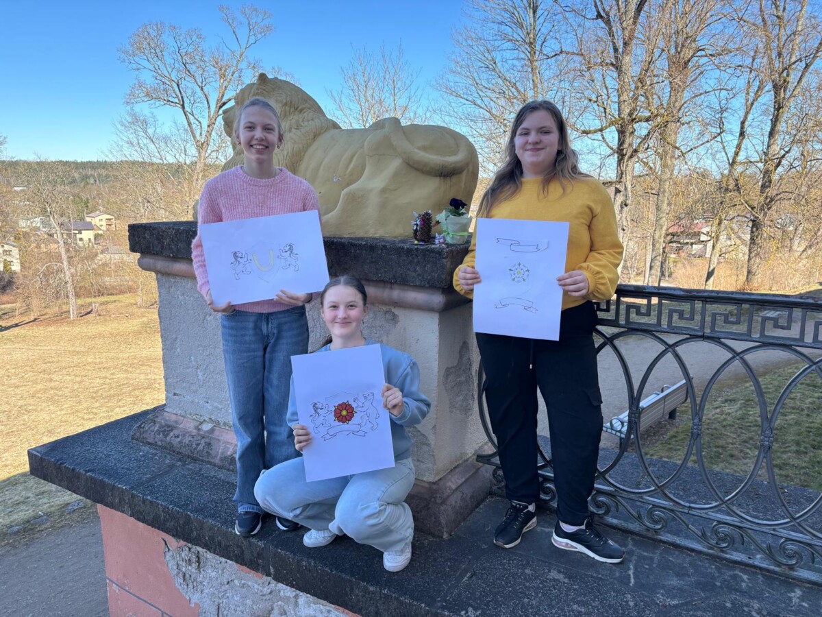 Trīs smaidīgas meitenes ārā tur rokās zīmējumus ar lentu un ziedu motīviem. Aiz viņām redzama zelta lauvas statuja, koki un tāla ainava saulainā dienā.